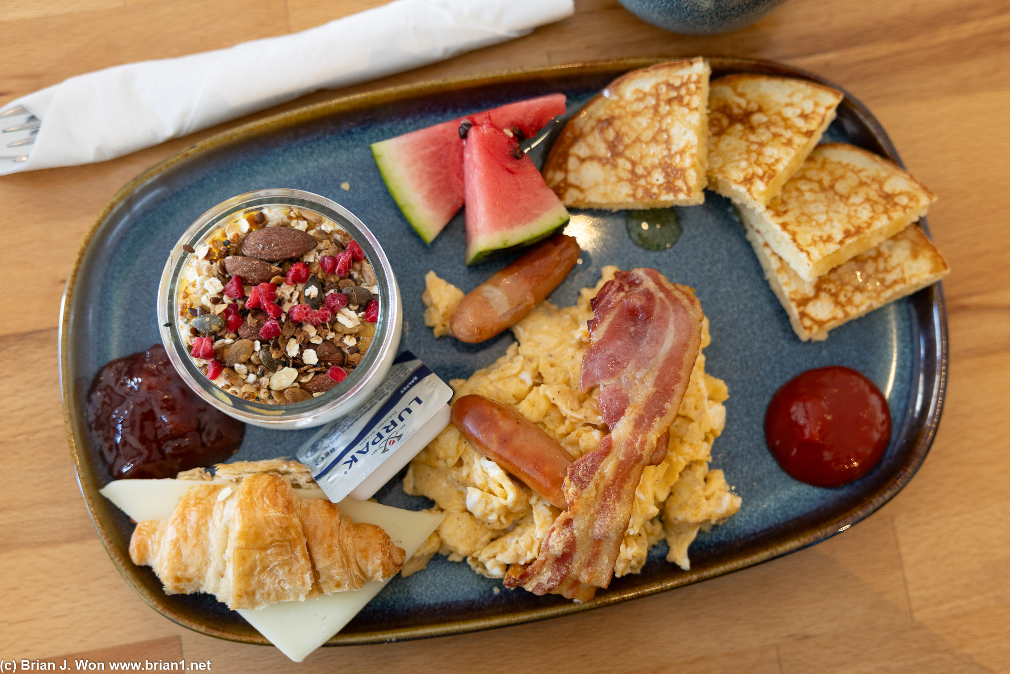 Breakfast with egg, bacon, sausage, cheese, croissant, watermelon, nuts, and yogurt.
